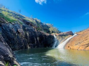 A photo of Serpentine Falls. The falls rush over orange-coloured rocks into a natural pool below. The sky above is clear and a brilliant blue colour.