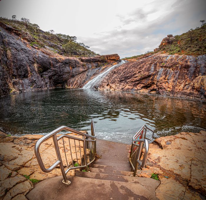A photo of stairs leading to the pool at the bottom of Serpentine Falls. The falls can be seen rushing over orange-coloured rocks in the distance. 