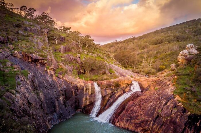 A photo taken by a drone of Serpentine Falls. Water rushes over orange-coloured rocks into a turquoise pool. Green trees and bushland surround the falls.