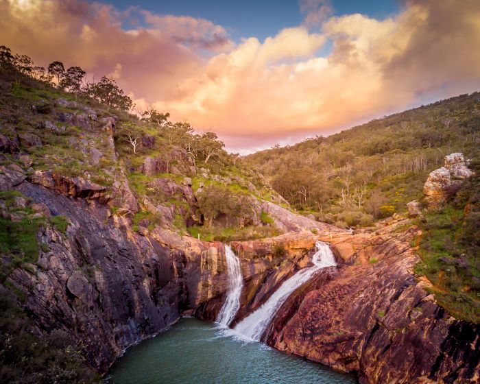 A photo taken by a drone of Serpentine Falls. Water rushes over orange-coloured rocks into a turquoise pool. Green trees and bushland surround the falls. 