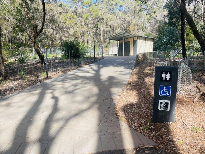 A photo of the path leading to the toilets at the picnic area of Serpentine National Park. The path is wide and flat, but on a slight incline. The toilet block can be seen in the distance and is surrounded by trees and bushland.