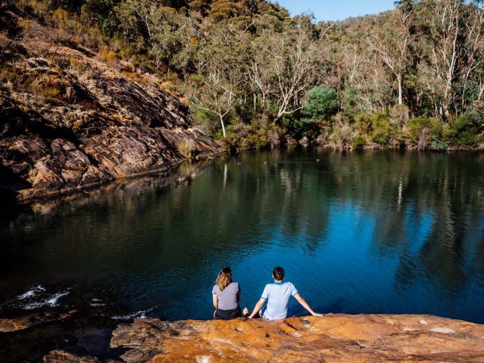 Two people sitting on a rock ledge on the edge of the natural pool at Serpentine Falls. The pool is a deep blue/green colour. Green trees and bushland can be seen in the distance.