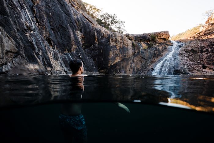 A photo of a person floating in the natural pool at the bottom of Serpentine Falls. The falls can be seen flowing over brown rocks in the background.