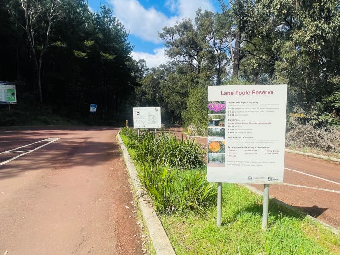 A photo of signs at the entry station of Lane Poole Reserve. The sign in focus explains the entry fees and campsite bookings. A road winds into the distance and is lined by tall trees.