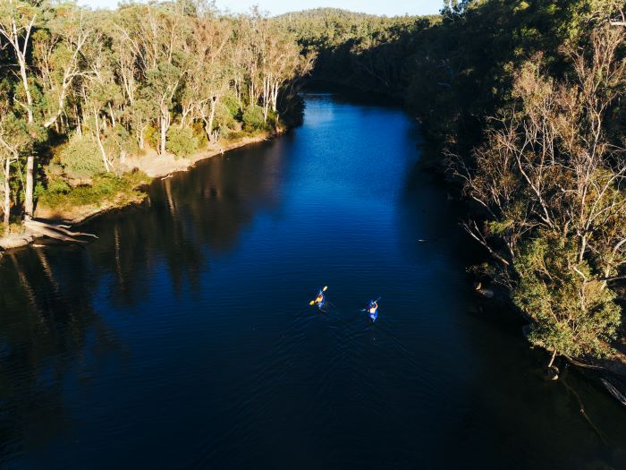 A photo taken from above by a drone, of two people kayaking on the river. The river is a dark blue colour and is lined by tall trees and bushland on both sides.
