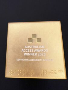 close up photograph of award. It is a gold cube with the wording - Australian Access Awards Winner 2023 Centre for Accessibility Australia - with braille beneath the wording.