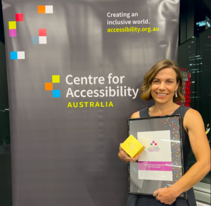 Dr Kelsie Prabawa-Sear stands in front of the Centre for Accessibility Australia banner, holding the awards for not-for-profit/community organisation app of the year, a framed certificate and a gold cube-shaped award.