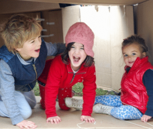 three children inside a large cardboard box, playing and calling out with happiness.
