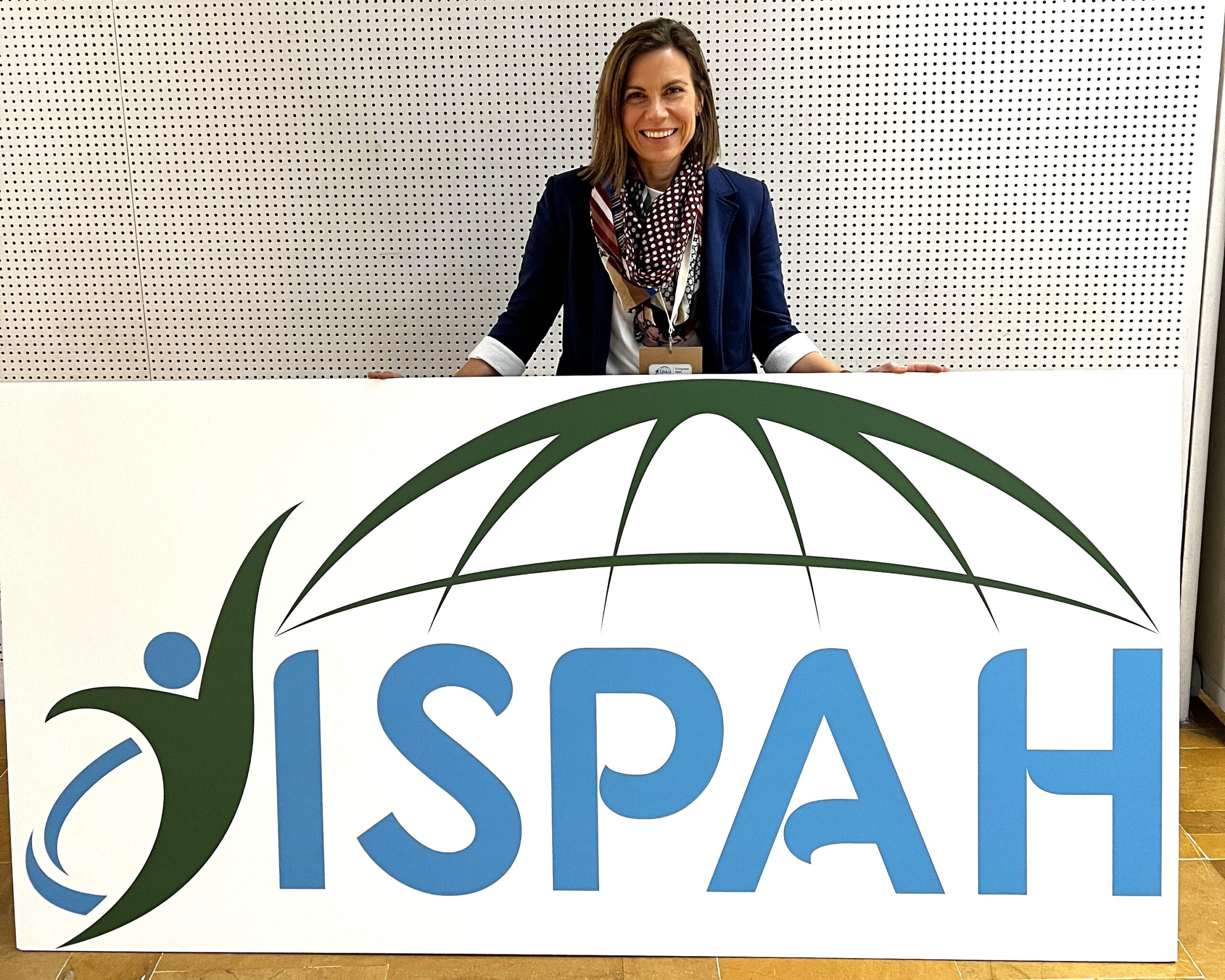 Nature Play WA CEO Dr Kelsie Prabawa-Sear stands behind a sign that says "ISPAH", which stands for International society for physical activity and health.