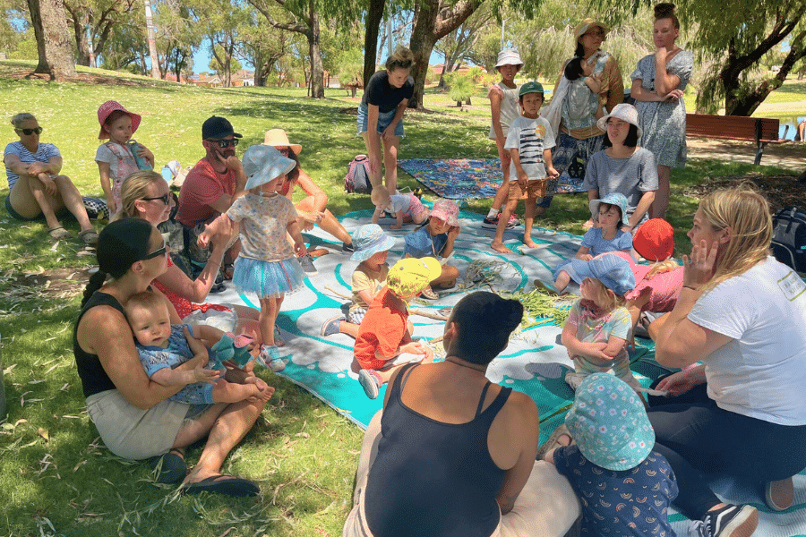 A group of adults and young children sit in a circle at a local park in the shade of surrounding trees. They are talking and taking part in a nature activity.
