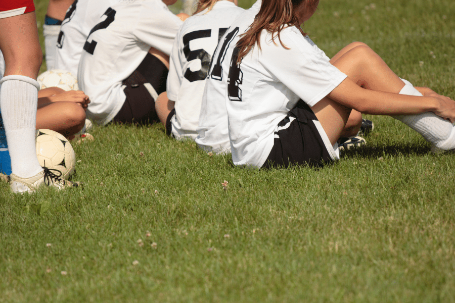 A group of girls sit on a field in soccer uniforms.