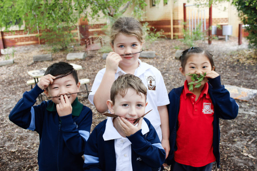 Primary school students outside smiling at the camera, holding sticks and leaves on their faces as moustaches.