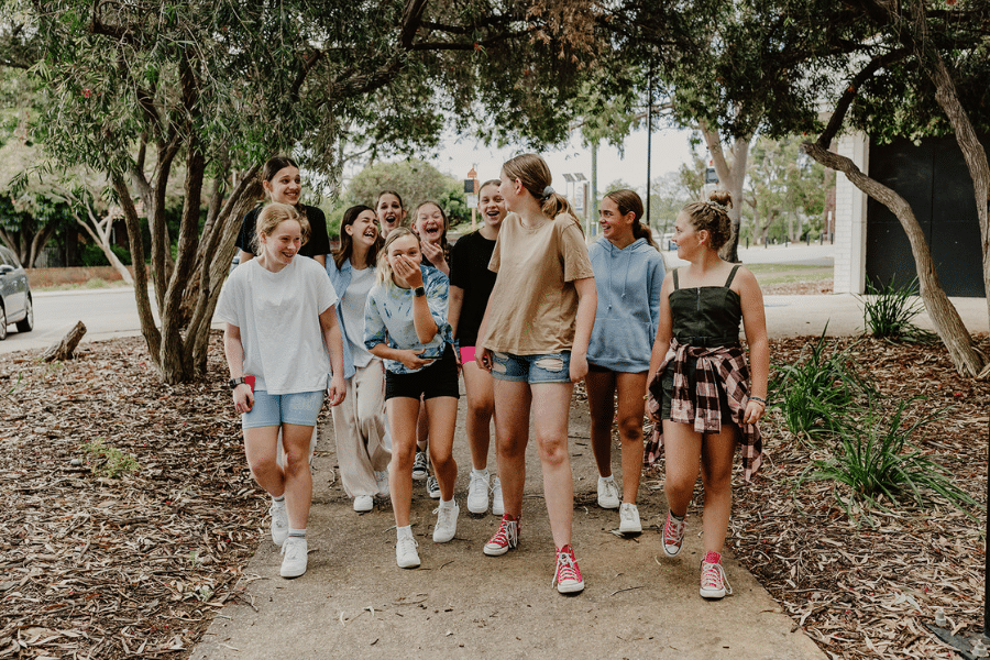 A group of tween and teen girls walk through a suburban park, laughing and chatting.
