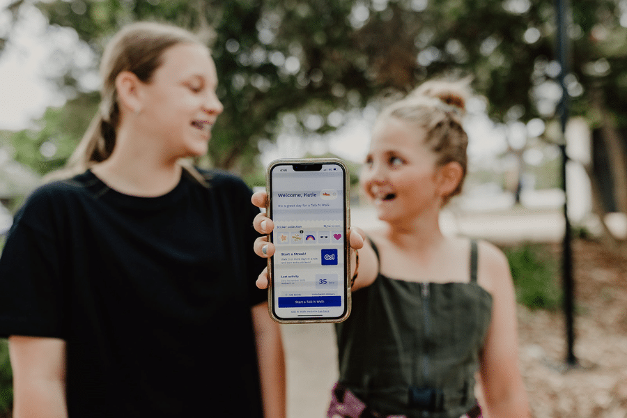 Two tween girls smile at each other while showing the Talk N Walk app on a phone screen to the camera.